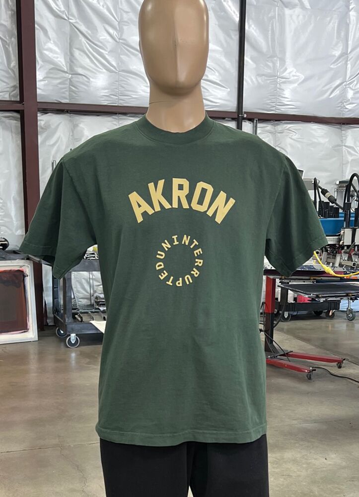 A mannequin is wearing one of the 100 Garment-Dyed Heavyweight Shirts - Text-Based Artwork with Vector Designs - Maximum of 2 Ink Colors - Soft Hand Screen Printing, featuring a green T-shirt with "AKRON" printed in large yellow letters across the front, inside a circular design. The background shows an industrial setting with machinery and workstations.