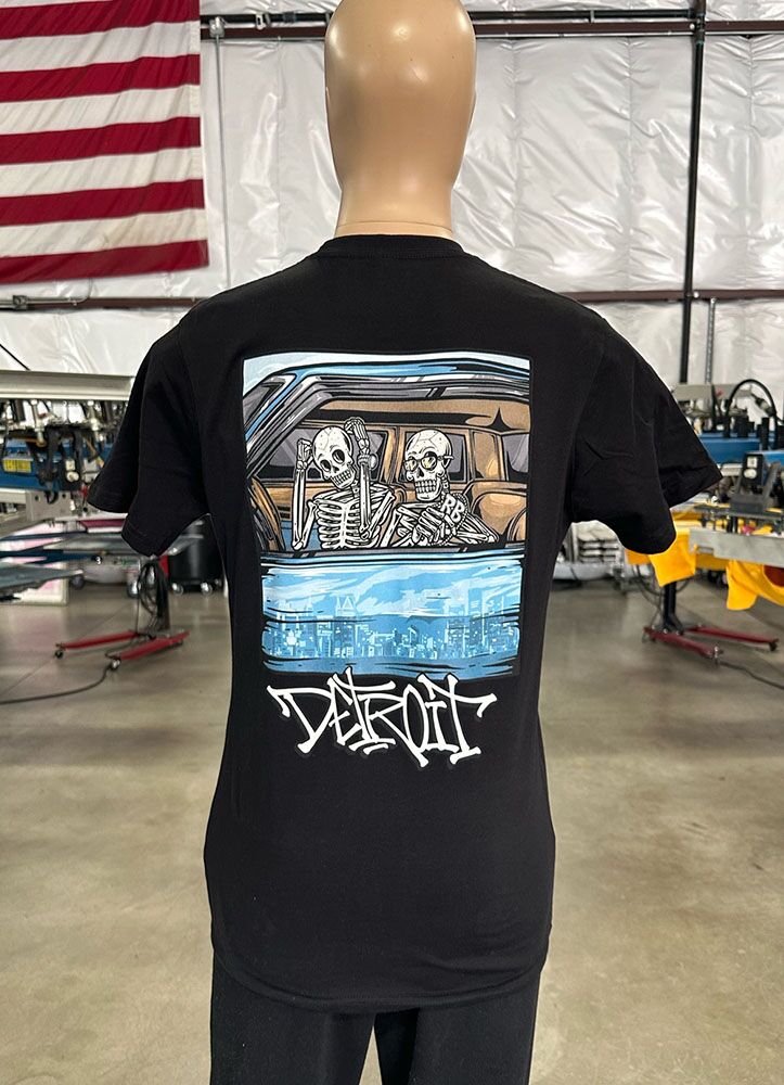 A mannequin displays a black 100 Digital Screen Printing- Garment-Dyed Heavyweight Shirt - Photo-Realistic Full-Color Printing - Hybrid Screen Print with Digital Color - 300 Resolution - Vivid Results with a graphic depicting two skeletons in a car, accompanied by the text "Detroit." An American flag is visible in the background.