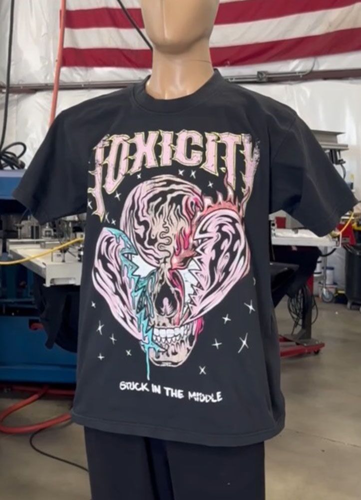 A mannequin displays a 100 Heavyweight Drop Shoulder Blanks - Streetwear - Maximum of 5 Ink Colors - Vibrant Loud Colors - Full Detail - Soft Hand Screen printing - Retail Ready with a colorful skull design and the word "TOXICITY" in bold letters, placed in an indoor setting with a U.S. flag partially visible in the background.