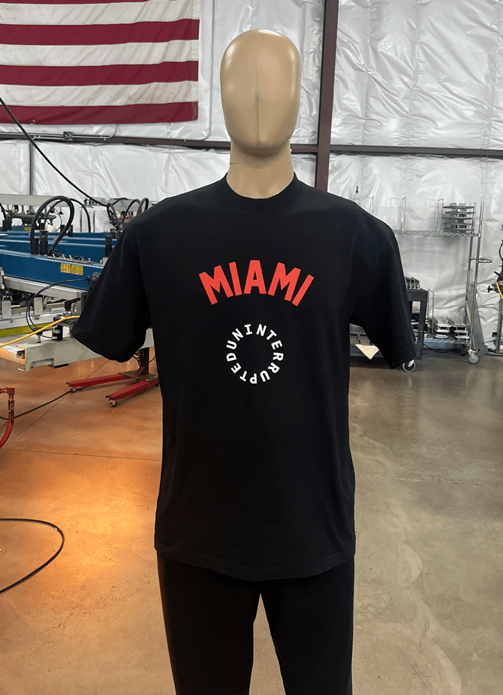 A mannequin displays one of the 100 Garment-Dyed Heavyweight Shirts - Text-Based Artwork with Vector Designs - Maximum of 2 Ink Colors - Soft Hand Screen Printing, featuring a black t-shirt with "MIAMI" in red text above and "INTERCEPTED" in white text below. The background includes an American flag and industrial equipment.